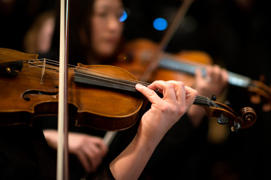 A close-up of a person's hand playing a violin, with blurred musicians in the background.