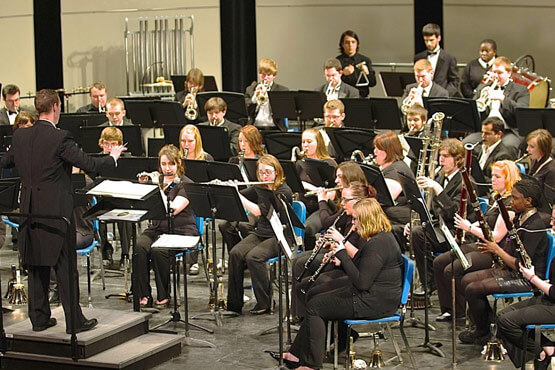 A conductor leads a large band performance with musicians playing various instruments on stage.
