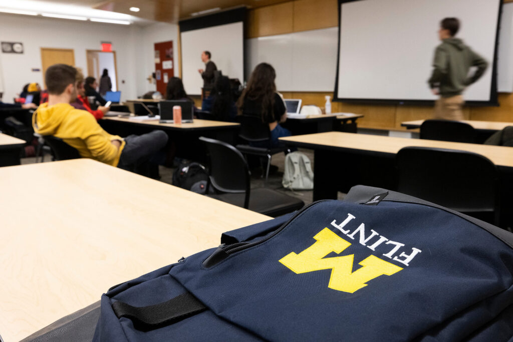 A classroom setting with students seated at desks, a backpack in the foreground displaying a logo, and a person speaking at the front.