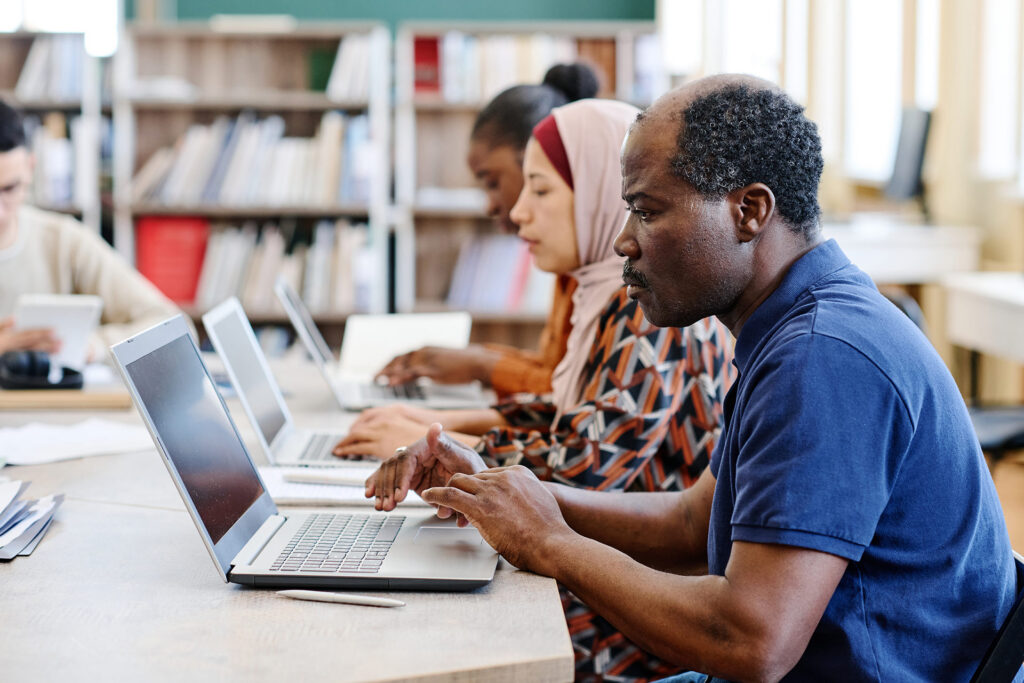 A group of people working on laptops at a table in a library, focused on their tasks.