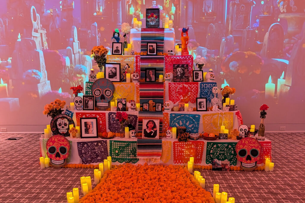 A colorful altar decorated with sugar skulls, flowers, candles, and framed photos, with marigold petals in the foreground.