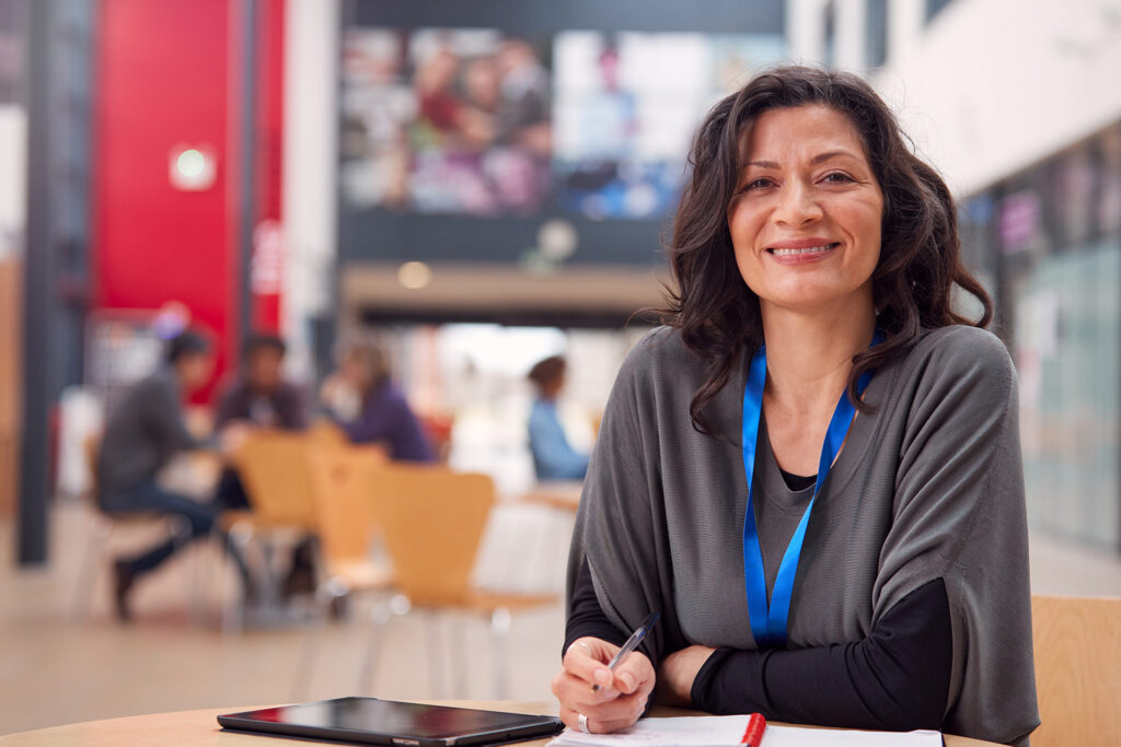 A person smiles while sitting at a table with a tablet and notebook, in a busy indoor space with others in the background.