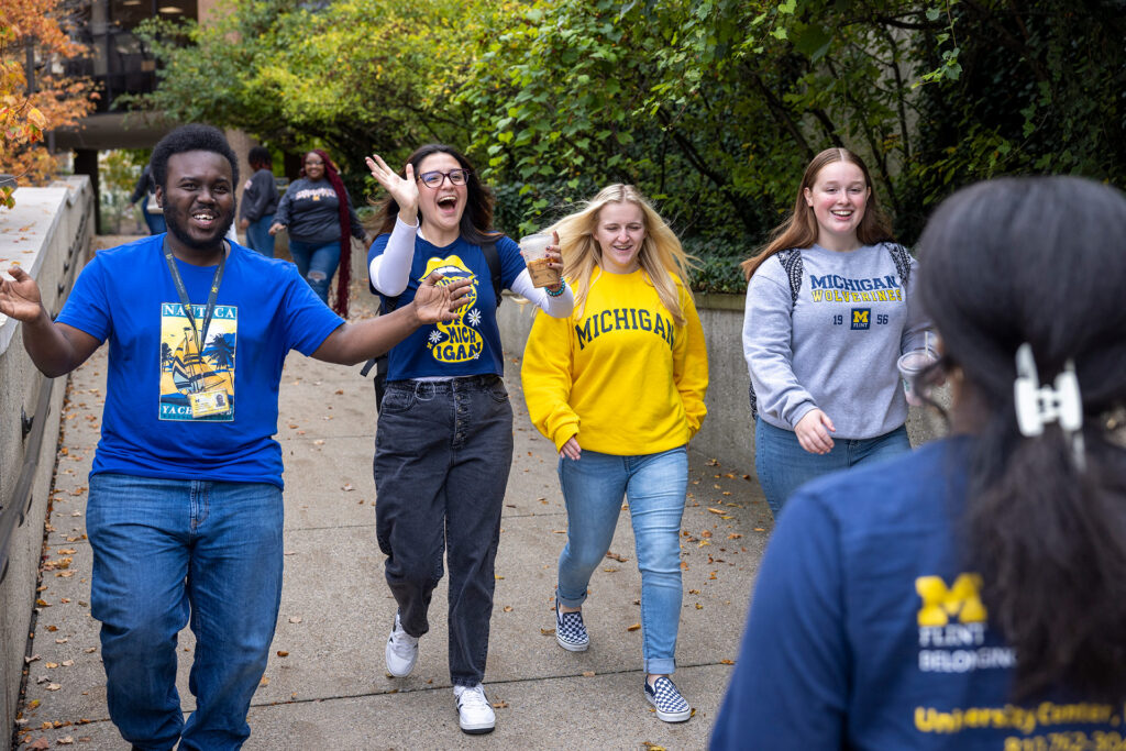 A group of four people walk joyfully along a path, wearing clothing with "Michigan" logos, surrounded by greenery.