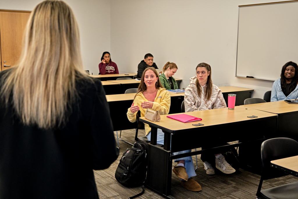 A person stands in front of a classroom, engaging with students seated at desks, some taking notes or listening.