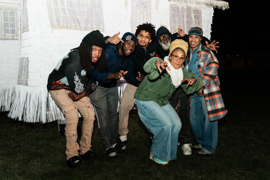 A group of six people poses playfully in front of a large, white, fringed structure at night.