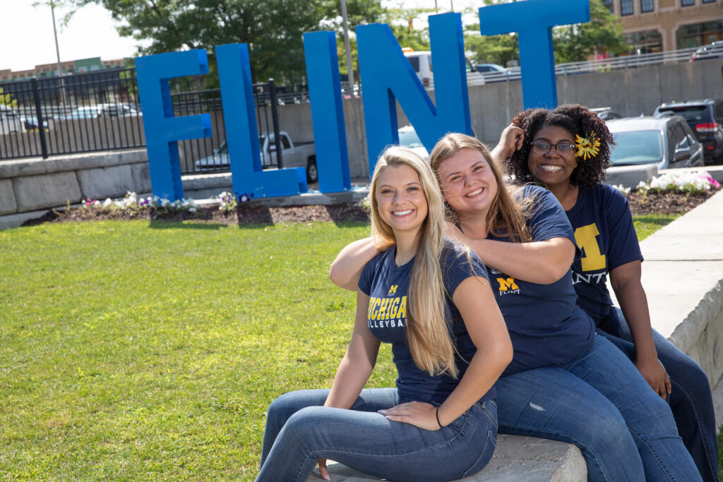 Three people sit on a stone ledge in front of a large blue sign that reads "FLINT," smiling and posing for the photo.