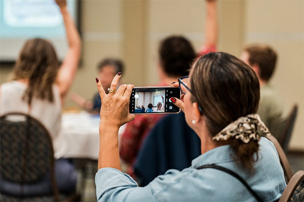 A person takes a photo with a smartphone while others raise their hands in a meeting or event setting.