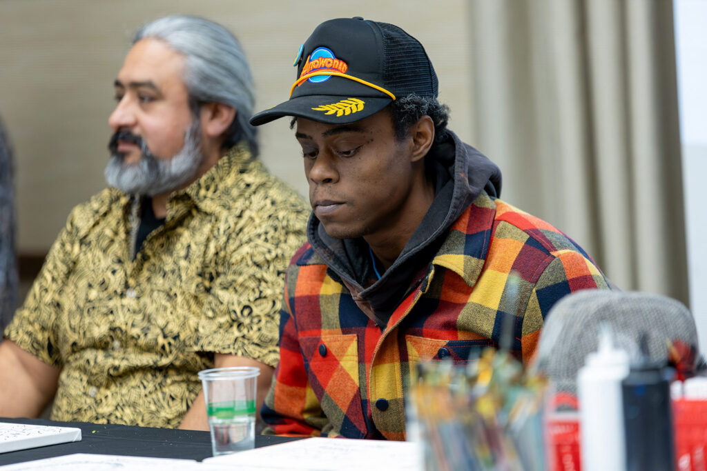 Two people are seated at a table, one wearing a patterned shirt and the other in a plaid jacket and cap, focused on their work.