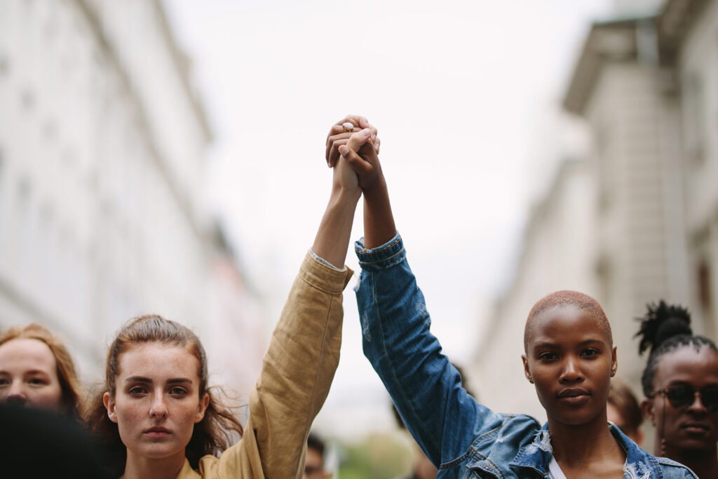 Two people hold hands high in the air during a protest, surrounded by a crowd in an urban setting.
