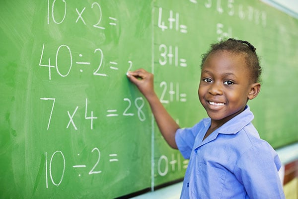 A person smiles while writing math equations on a green chalkboard in a classroom setting.