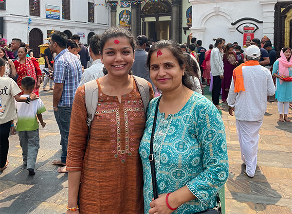 A person and a woman stand together, smiling, in a crowded outdoor setting with people and buildings in the background.
