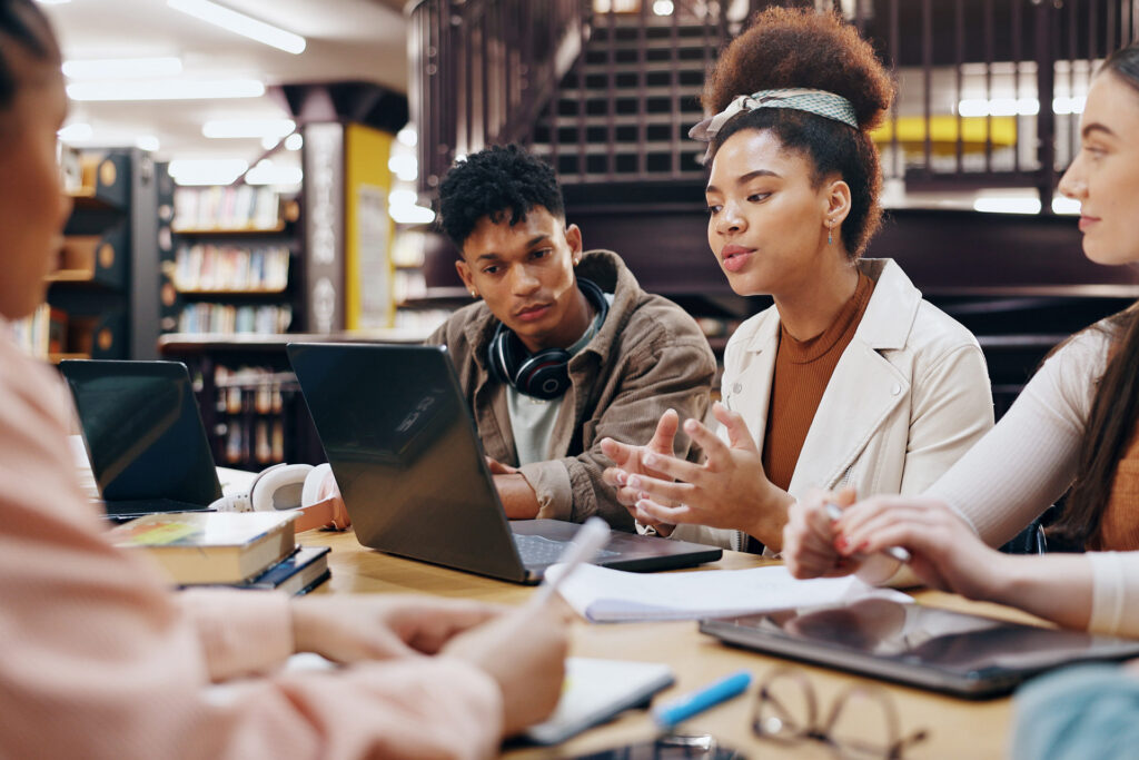 A group of four people engaged in a discussion at a table in a library, with laptops and books in front of them.