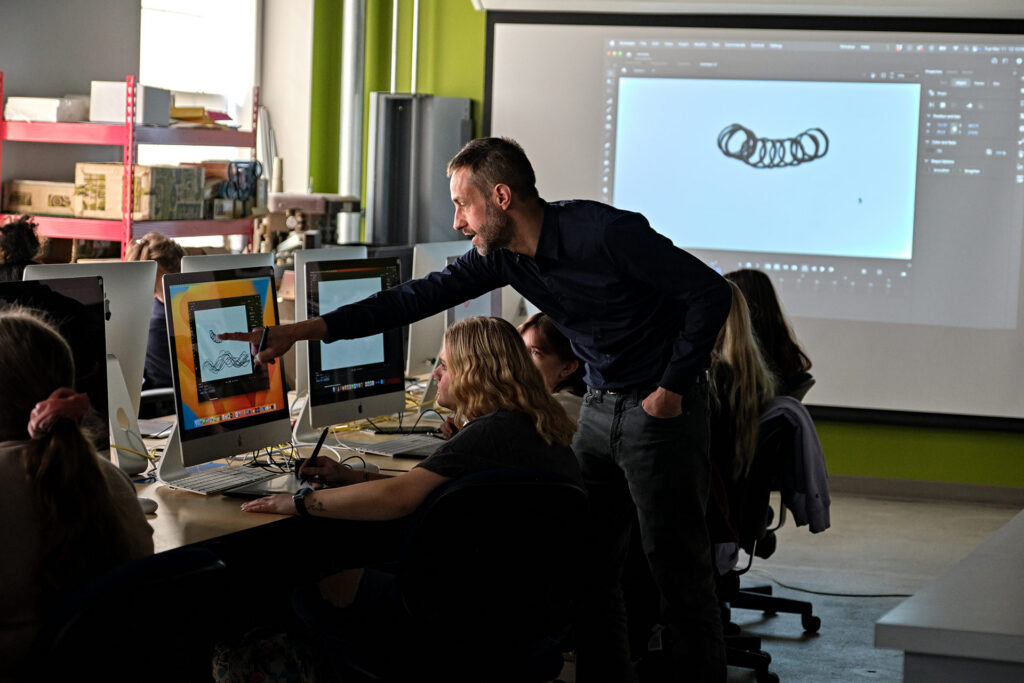 A person points at a computer screen while teaching a group of students in a classroom with multiple computers.