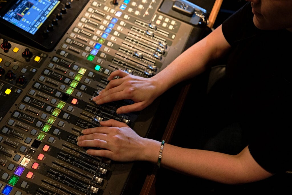 A person adjusts audio levels on a mixing console with multiple sliders and colorful buttons.