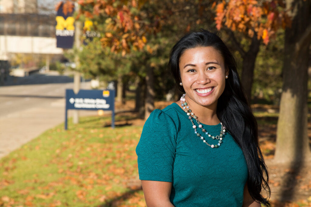 A person smiles while standing outdoors in front of a sign for the University of Michigan, surrounded by autumn foliage.