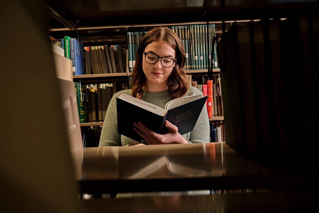 A person reading a book in a library, viewed through the shelves of books.