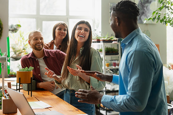 A group of four people are engaged in a lively conversation in a bright, plant-filled space.