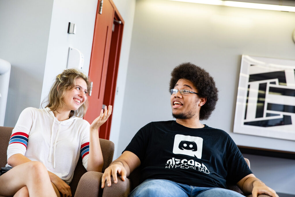 Two people are sitting in a casual setting, smiling and engaging in conversation.