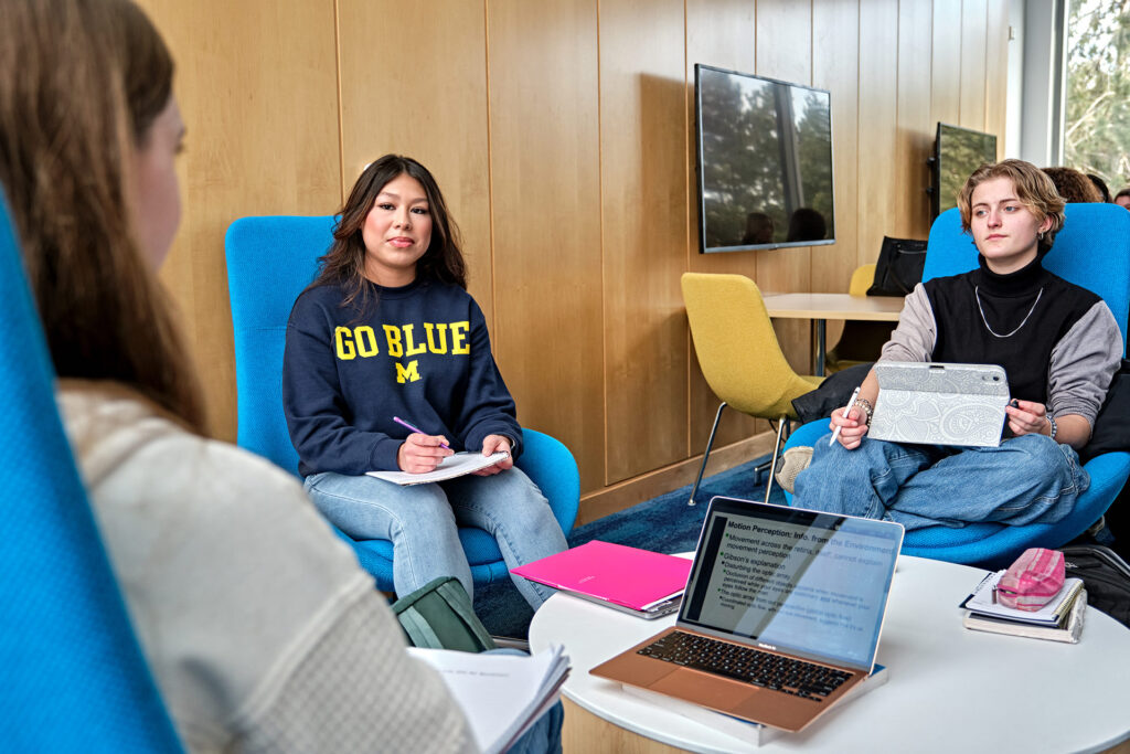 Three people are seated in a study area, engaged in discussion, with a laptop and notebooks on a table in front of them.