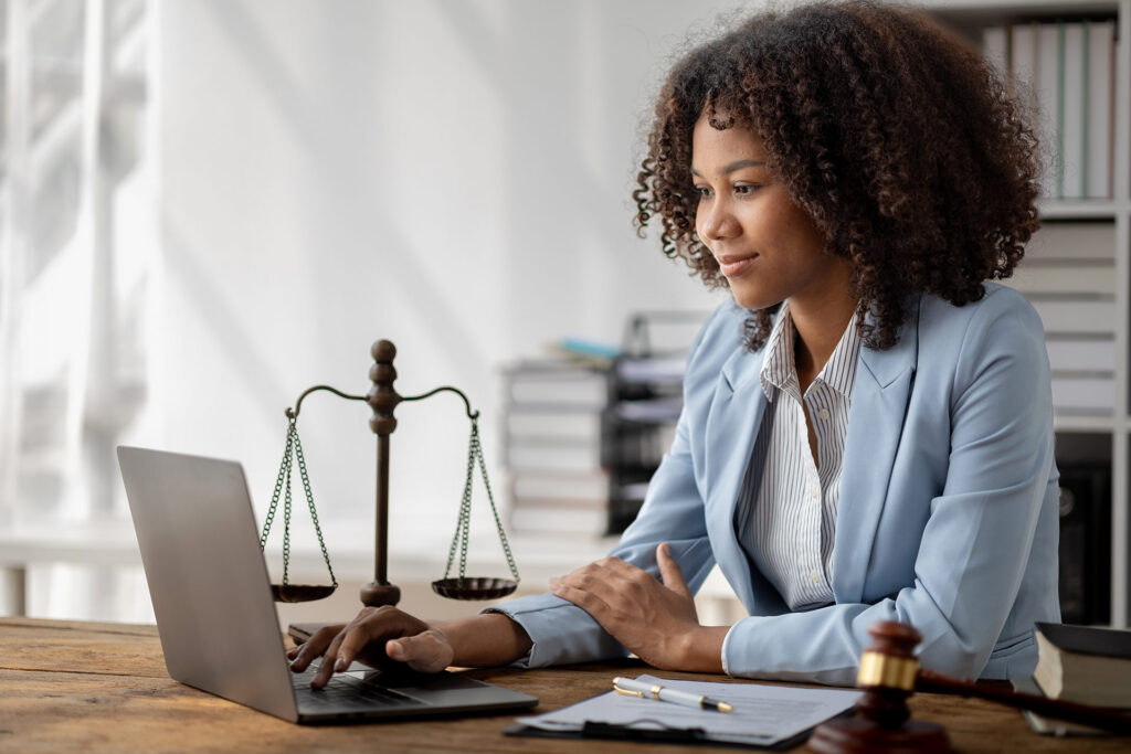 A person in a light blue suit is working on a laptop at a desk with a gavel and a scale of justice nearby.