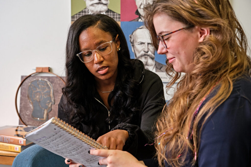 Two people are sitting together, discussing notes in a notebook, with educational posters in the background.