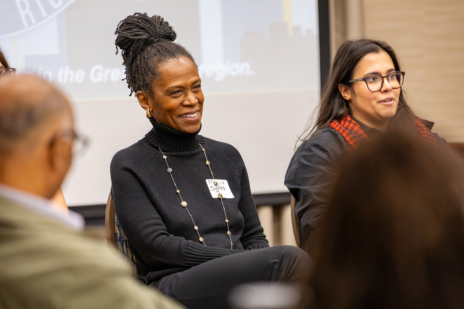 A person smiles while seated on a panel, with another person speaking beside them, in a conference setting.