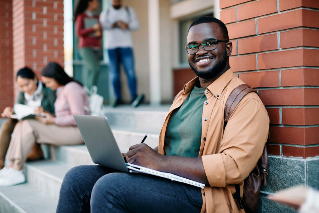A person sits on steps with a laptop, smiling and writing, while others study in the background.