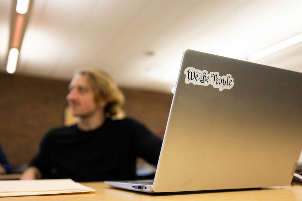 A laptop with a "We the People" sticker is in focus, while a person is blurred in the background.