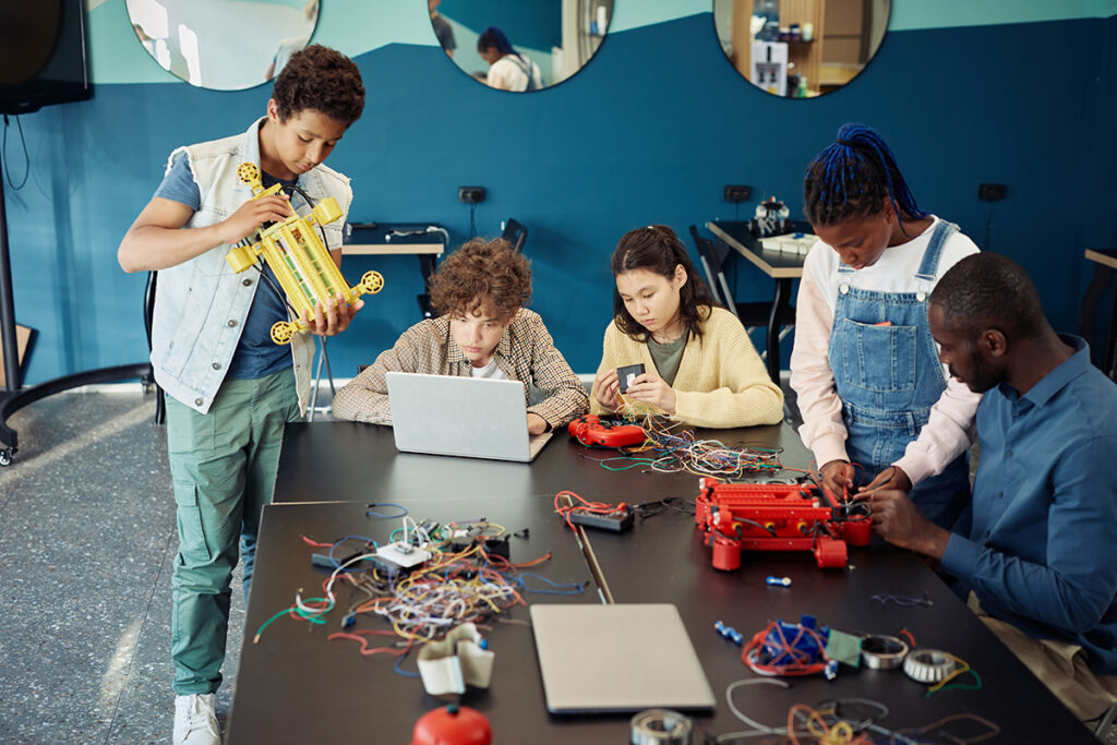 A group of four people work on various projects at a table, with one person holding a toy car and others using a laptop and phone.