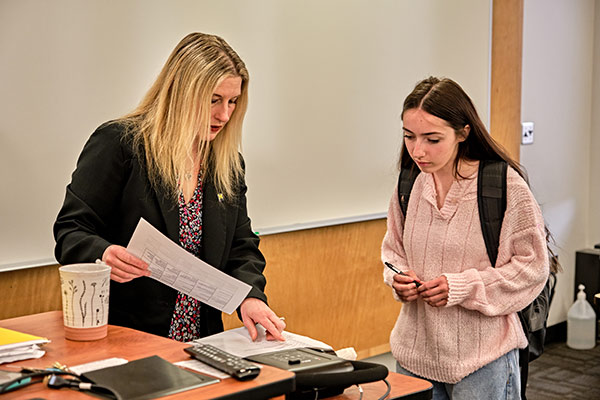 A person hands a sheet of paper to another person in a classroom setting, with a desk and various items visible.