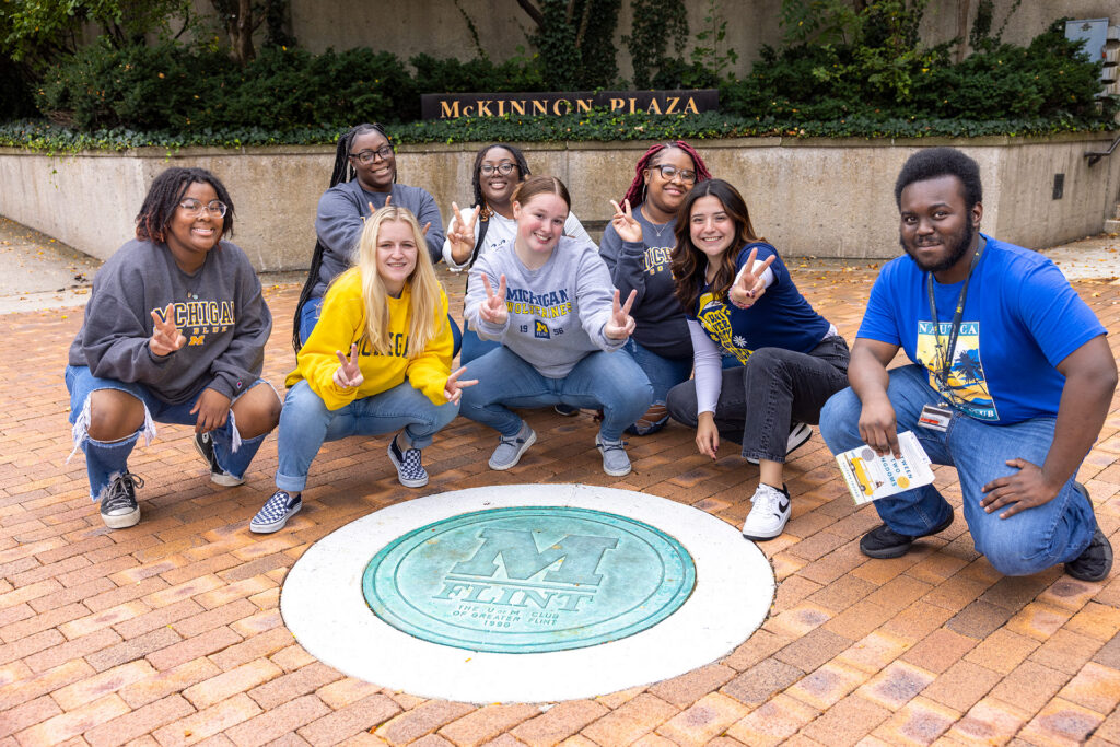 A group of seven people poses around a circular emblem on the ground at McKinnon Plaza, smiling and making peace signs.