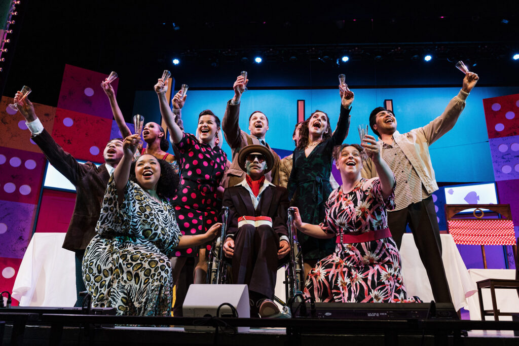 A group of ten performers joyfully pose on stage, raising glasses in celebration against a colorful backdrop.