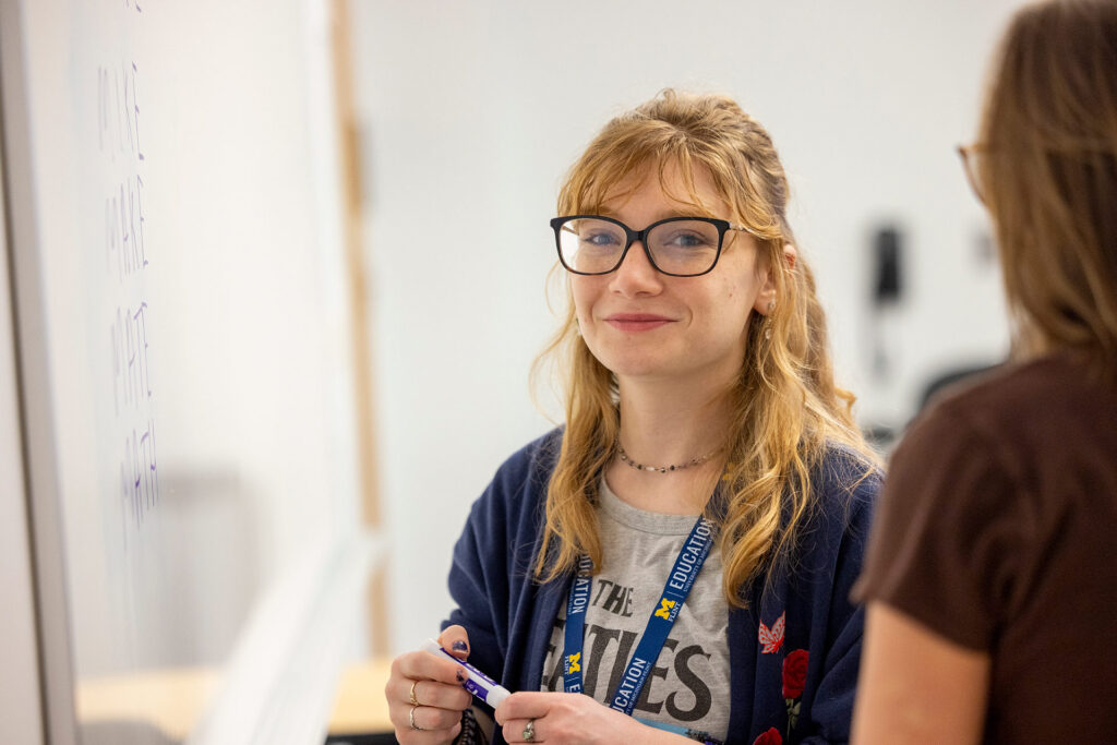 A person smiles while holding a marker, standing near a whiteboard with writing, engaged in conversation with another person.