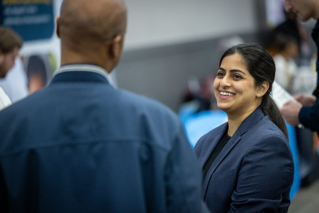 A person in a suit smiles while engaging in conversation with another person, whose back is facing the camera.