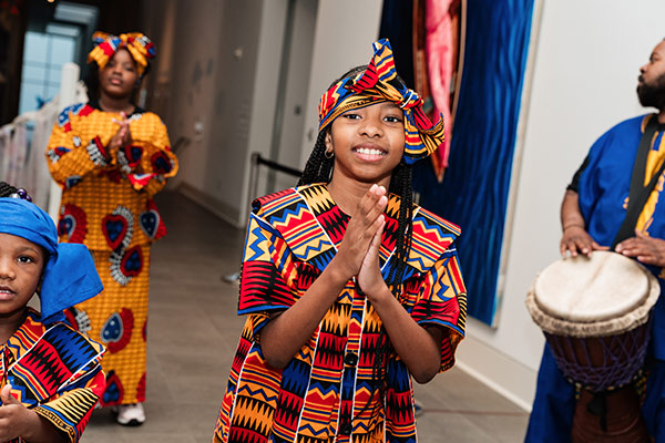 A group of children in colorful traditional attire perform, with one child smiling and clapping hands in a gallery setting.