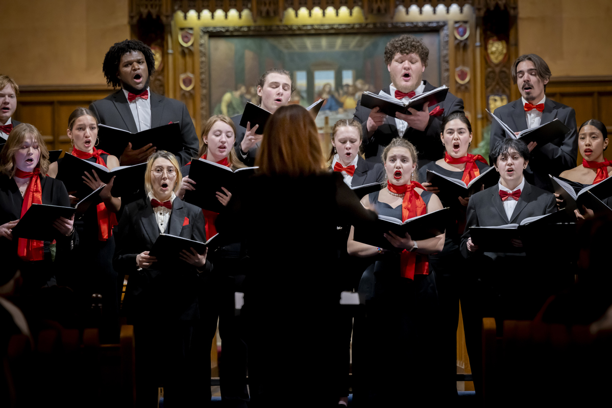 A choir of singers in formal attire performs with sheet music, directed by a conductor in front of a decorative backdrop.