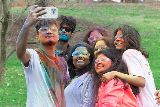 Students covered in colorful powder taking a group selfie outdoors during a Holi celebration.