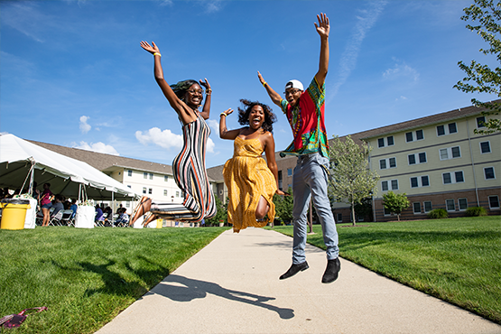 Three students jumping in the air on a sunny walkway outside the campus residence halls.