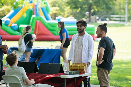 Students talking around a display table at an outdoor campus event with an inflatable bounce house behind them.
