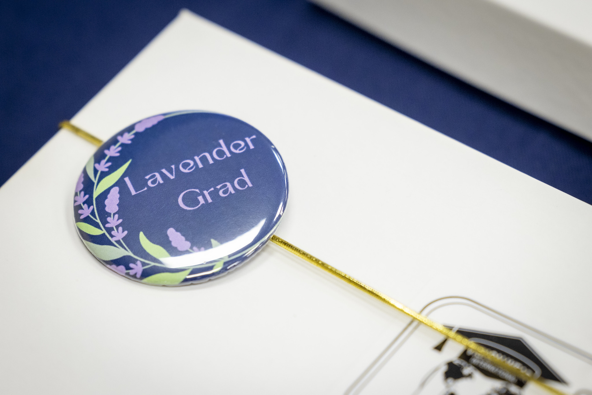 A button with the text "Lavender Grad" surrounded by lavender flowers, attached to a white card with a gold string.