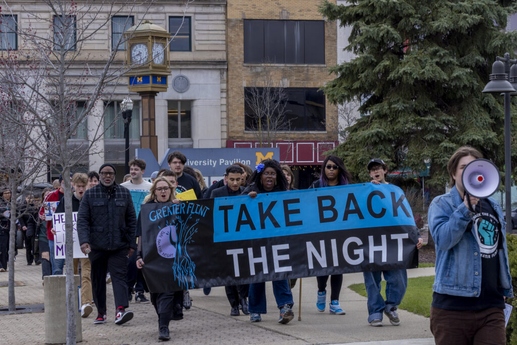 A group of people marches holding a banner that reads "TAKE BACK THE NIGHT," with a clock tower in the background.