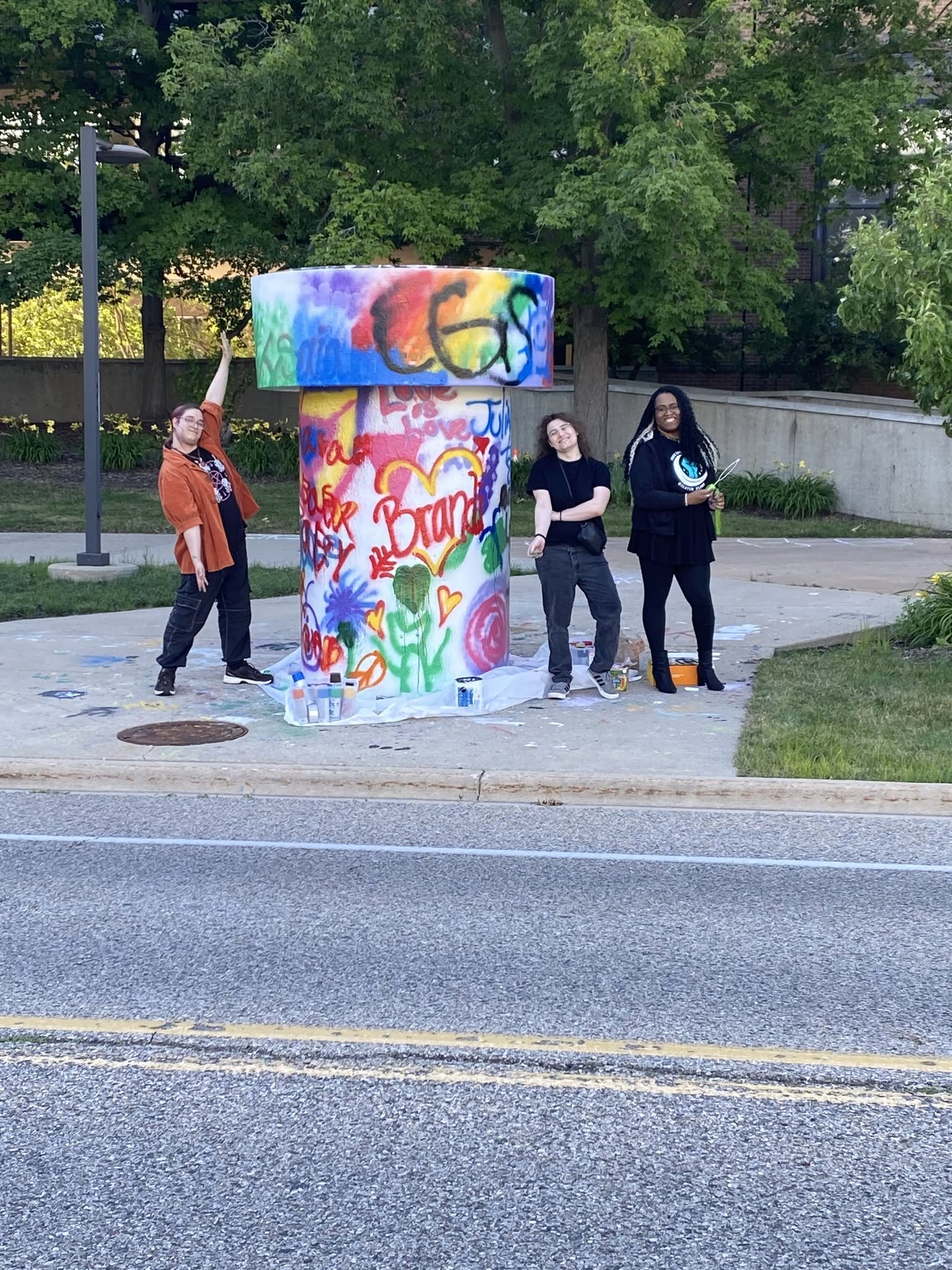 Three people pose next to a colorful, graffiti-covered pillar in a park setting, with paint supplies scattered nearby.