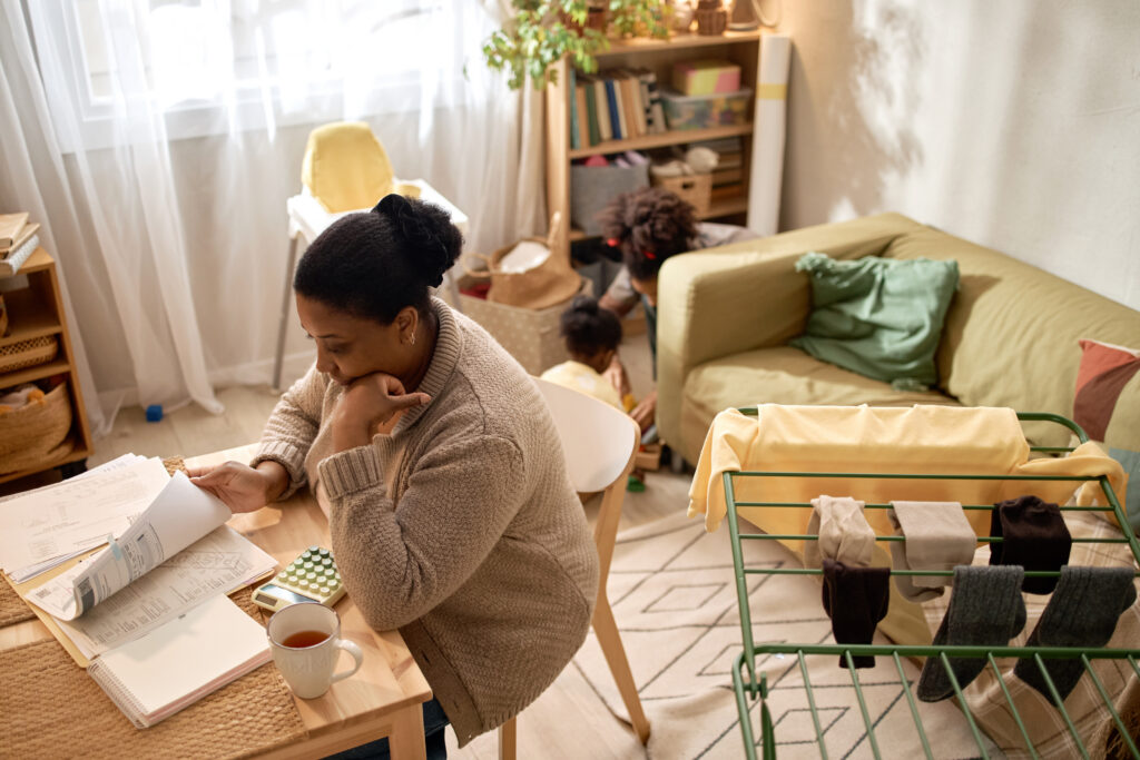 A person sits at a table reviewing documents with a cup of tea, while a child plays on the floor nearby and laundry dries on a rack.
