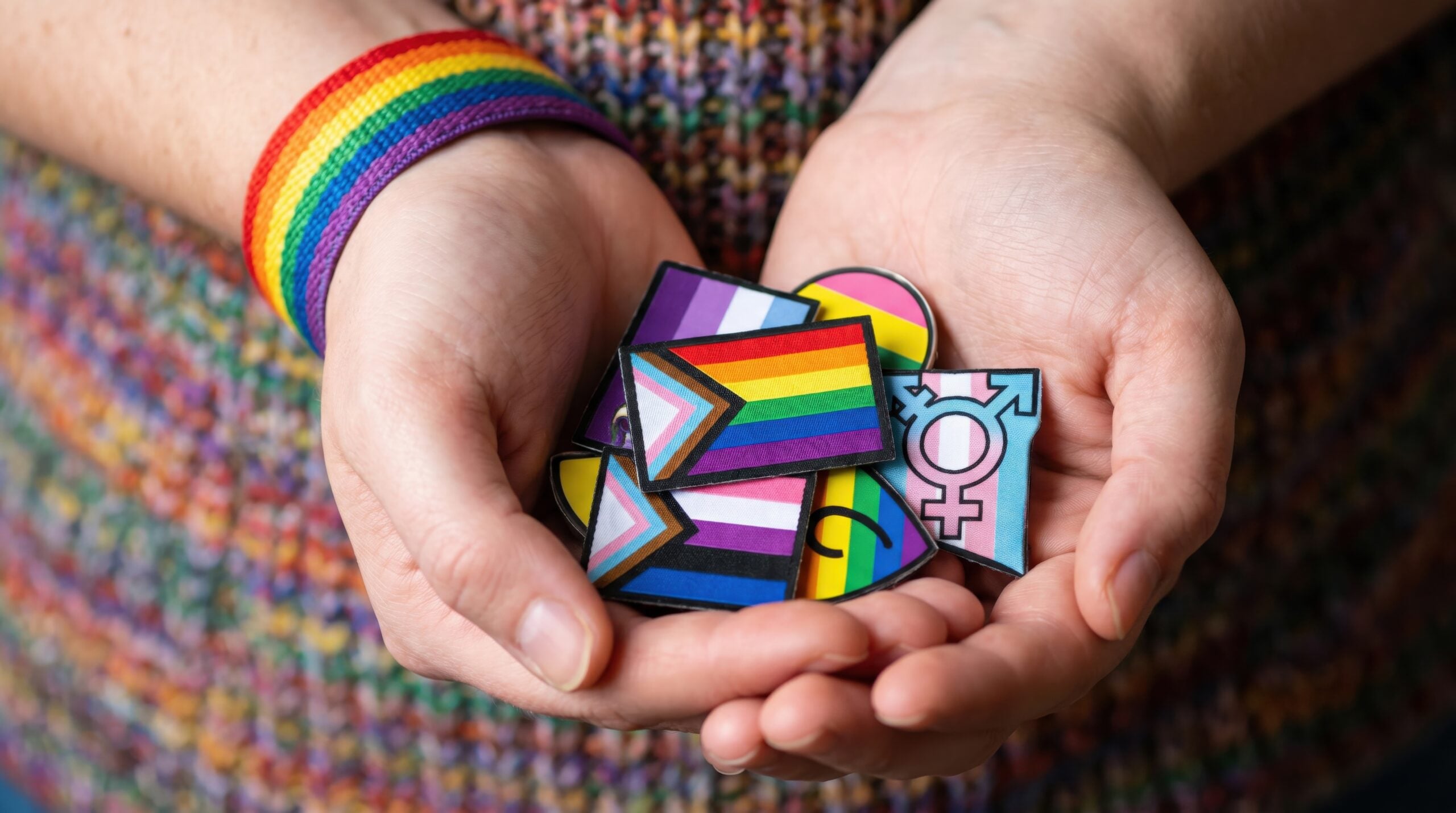 A person holds a collection of colorful LGBTQ+ pride pins in their hands, wearing a rainbow bracelet.