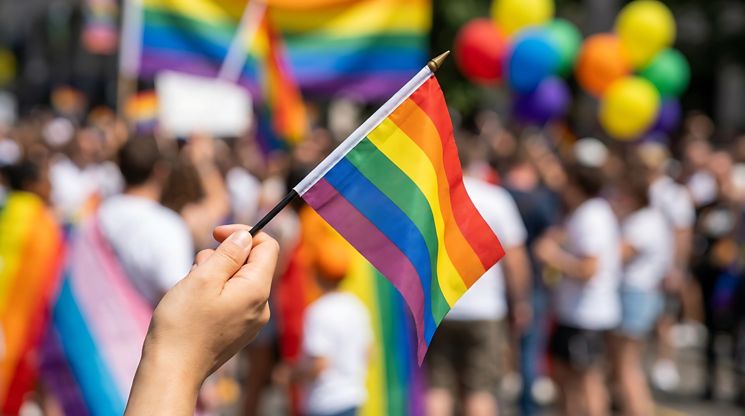 A hand holds a small rainbow flag in a crowd celebrating, with colorful balloons and larger flags in the background.