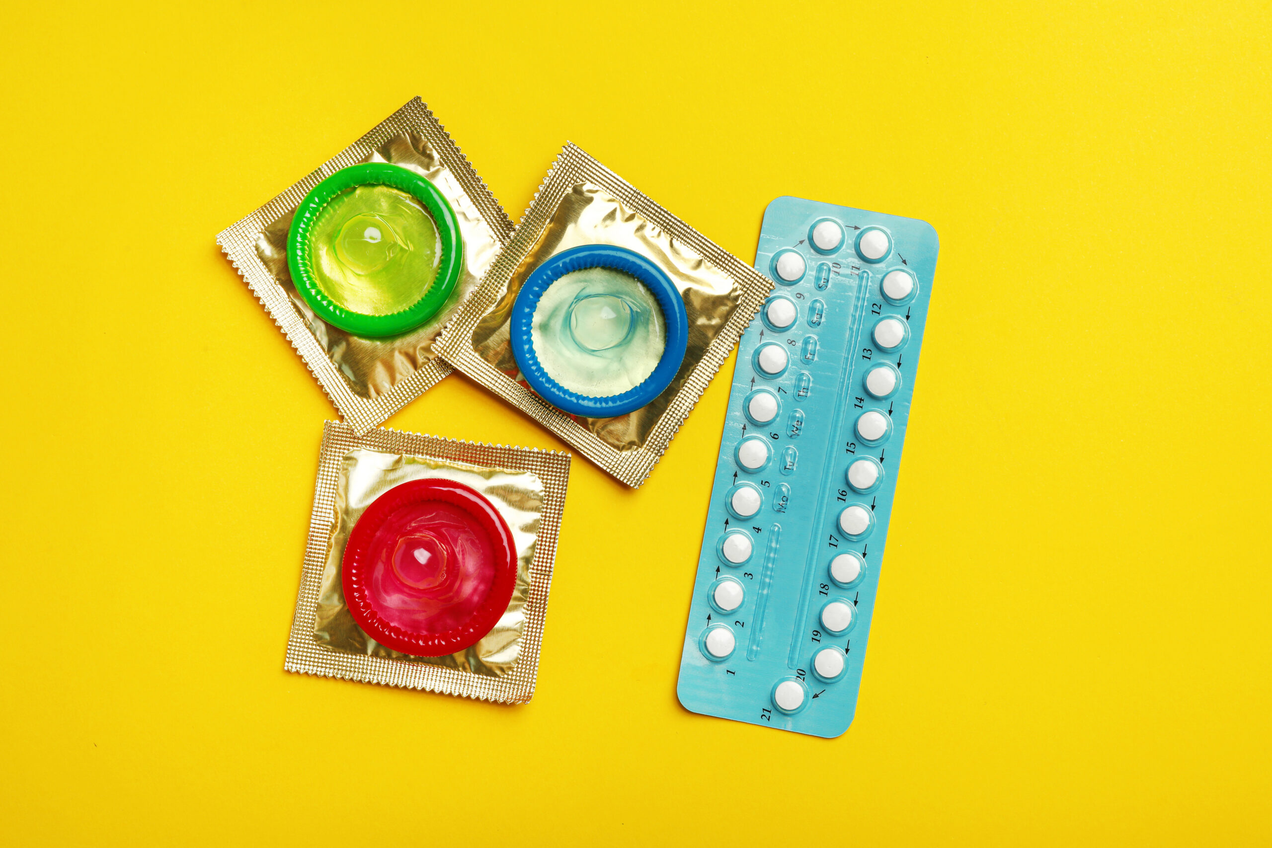 Three colored condoms and a pack of birth control pills are arranged on a bright yellow background.
