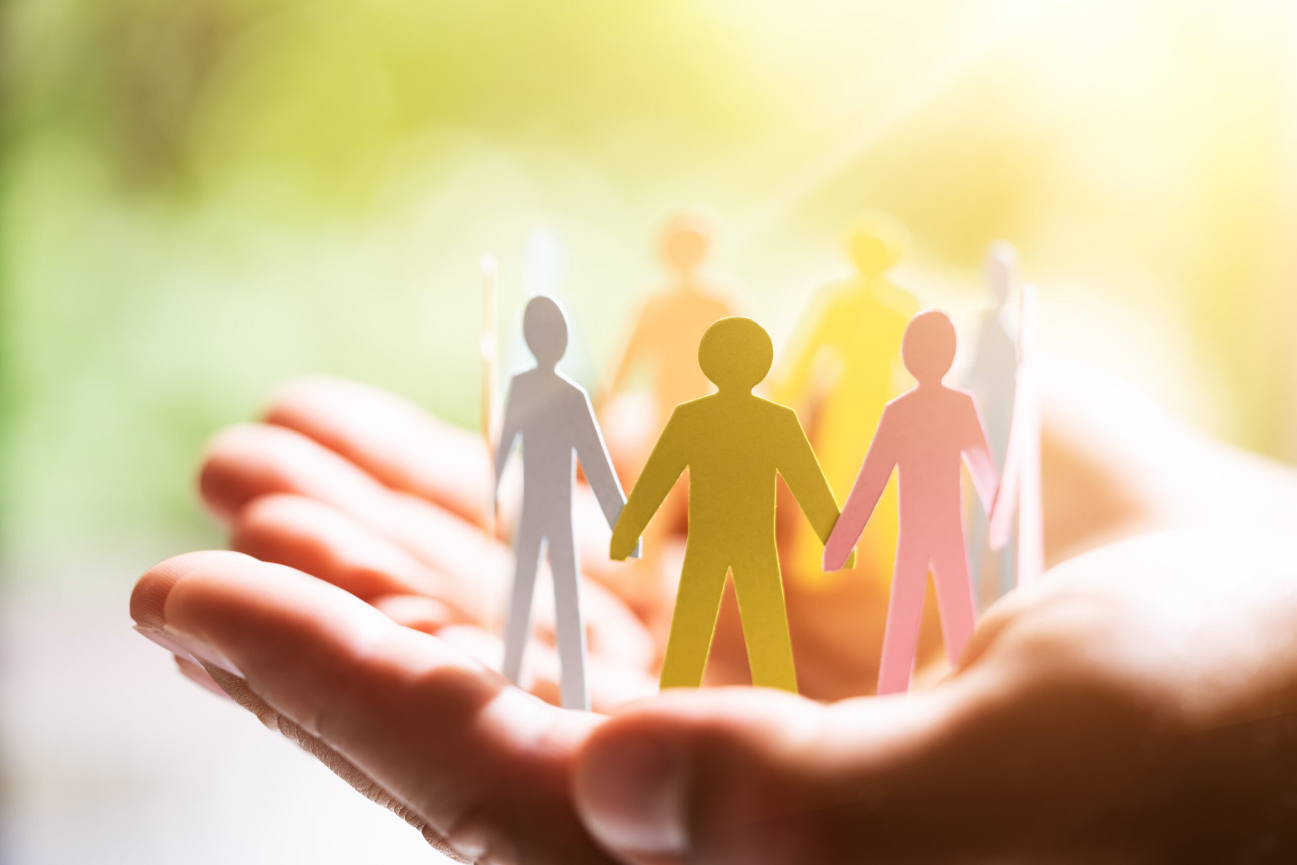 A person's hands hold colorful paper cutouts of people, symbolizing community and support, with a soft background light.