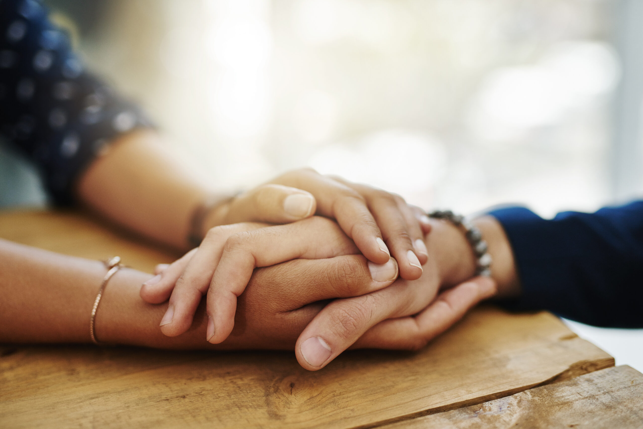 Two hands are gently resting on top of each other on a wooden table, conveying a sense of connection and support.