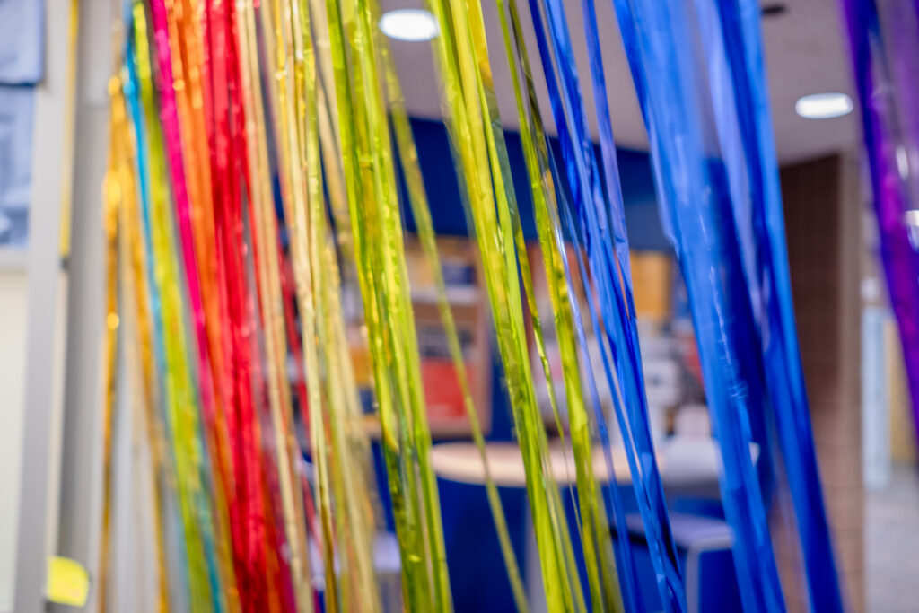 Colorful streamers in red, pink, yellow, green, blue, and purple hang vertically in a bright indoor setting.