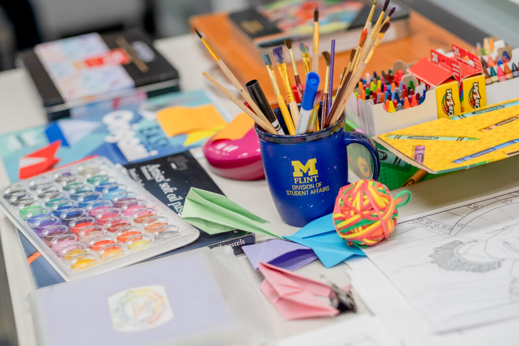 A cluttered table with art supplies, including a mug with "Flint Division of Student Affairs," paint palettes, paper, and crayons.